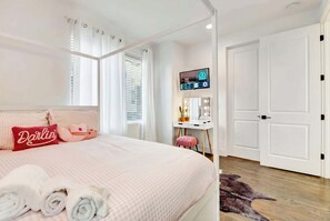 A neatly made bed with pink bedding and a red pillow, a small desk with a mirror and a TV, and a white door in a bright room. - Nashville bachelorette house with hot tub, karaoke stage & game room, sleeps 13 (Nashville)