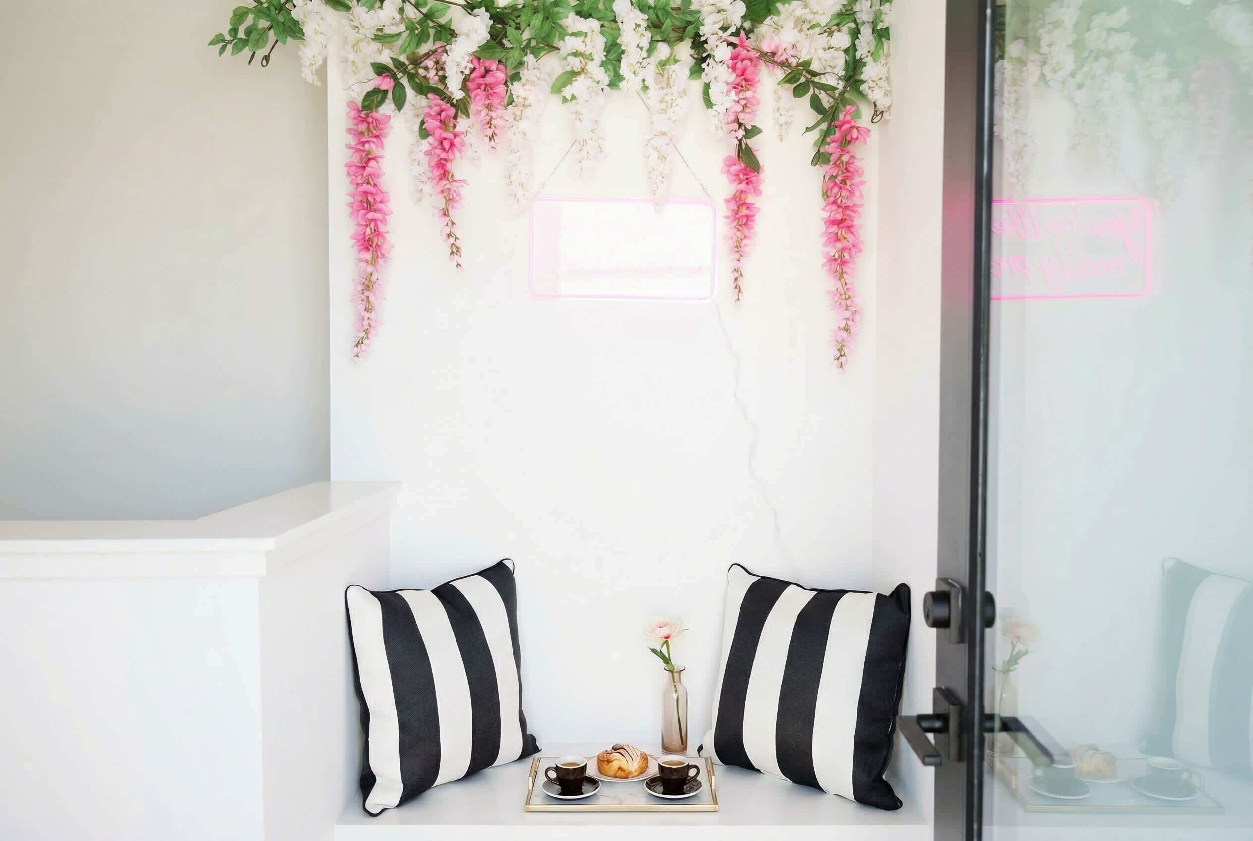 A room with a white wall, black and white striped pillows, a small table with pastries and a vase of flowers, and hanging floral decorations.