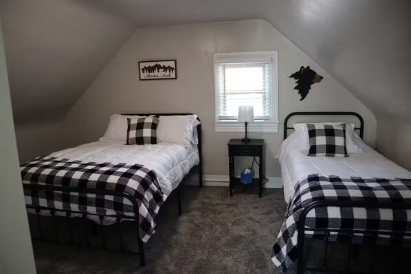 A small, triangular room with two beds, each with a checkered blanket, a nightstand with a lamp, a window with blinds, and a wall-mounted decoration. - Mount Powell Getaway walking distance to downtown (Deer Lodge)