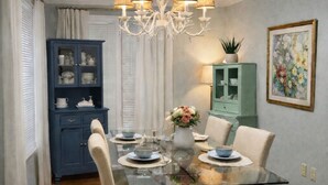 A dining room with a glass table, beige chairs, a floral rug, a chandelier, and blue cabinets.