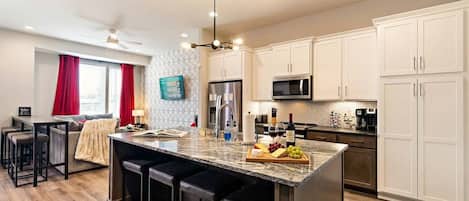 Una cocina moderna con una isla central, taburetes de bar, electrodomésticos de acero inoxidable y un comedor con sofá y sillas.