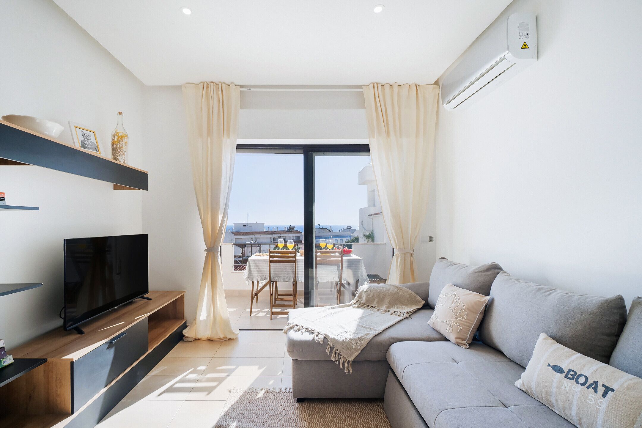 Ein modernes Wohnzimmer mit Fernseher, grauem Sofa und einem Balkon mit Tisch und Stühlen.