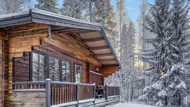 Puinen mökki, jossa on kuisti ja suuret ikkunat, ja jonka ympärillä on lumipeitteisiä puita.