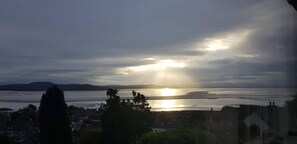 A coastal view with a sunset over water, buildings, and a prominent tree in the foreground. - Detached house with three flats | Amazing views (Grange-over-Sands)