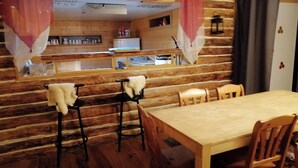 Un interior rústico de madera con una mesa de comedor y sillas, una zona de cocina con refrigerador, y una ventana con cortinas.