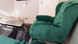 A green armchair in a room with a flat-screen TV mounted on the wall, a white door, and a patterned rug. - KARMİLİFE KONAKLAMA (Karaman)