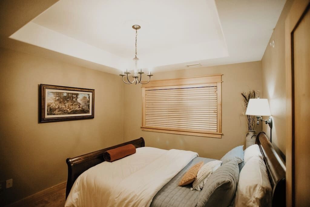 Un dormitorio con cama, candelabro, ventana con persianas, un cuadro en la pared y lámpara de noche.