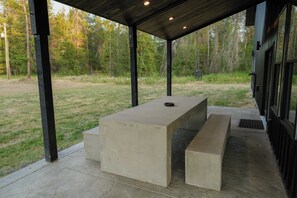 Une terrasse extérieure couverte, avec une table et des bancs en béton, entourée d’une pelouse et d’arbres.