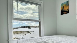 Un dormitorio con un amplio ventanal que da a una playa y un cuadro de un faro.