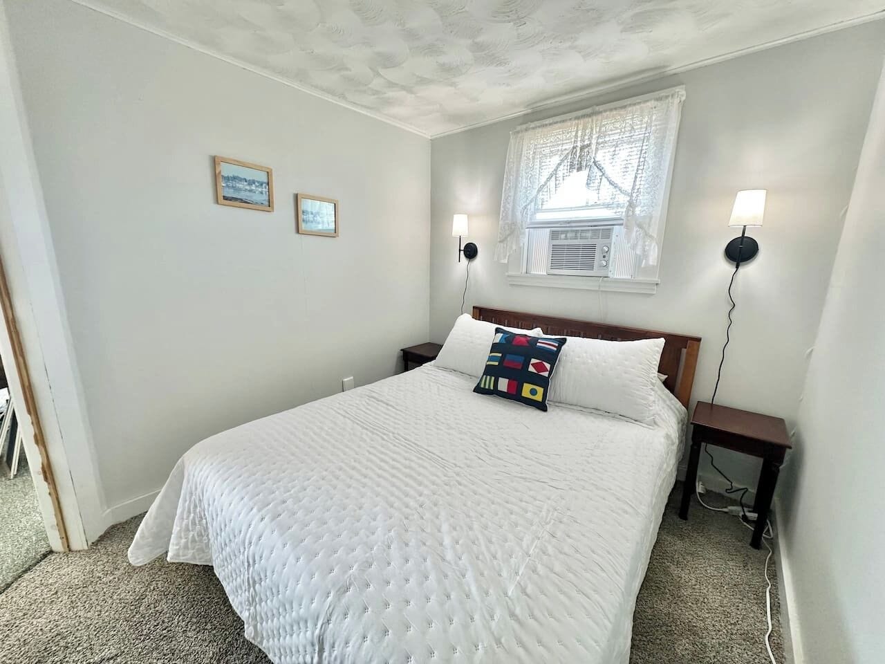 A bedroom with a large bed, two bedside tables, a window with lace curtains, and two wall-mounted lamps.