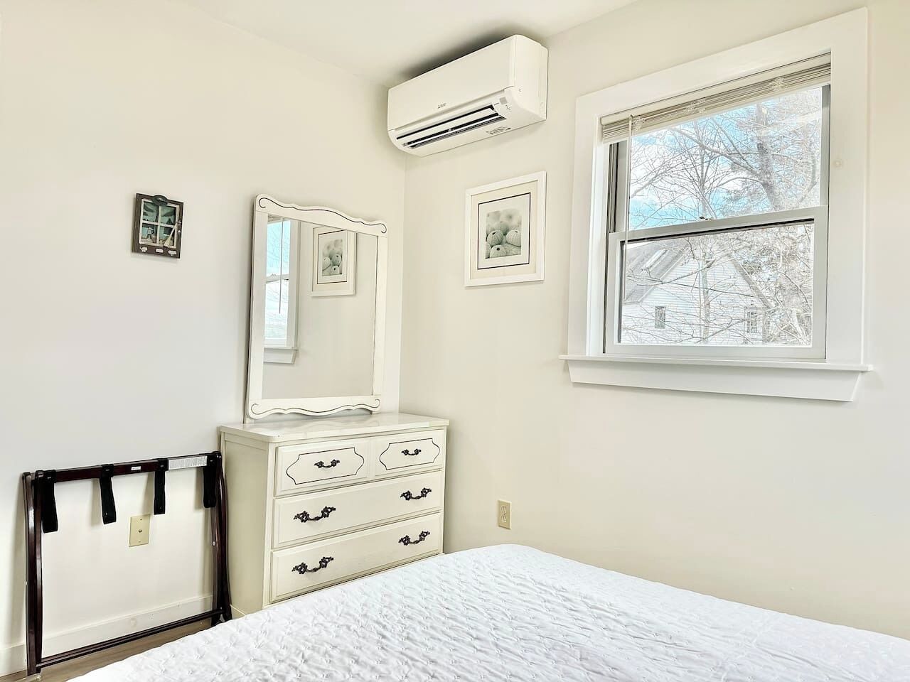 Un dormitorio con cama, cómoda, espejo, equipo de aire acondicionado y ventana con vista a árboles.