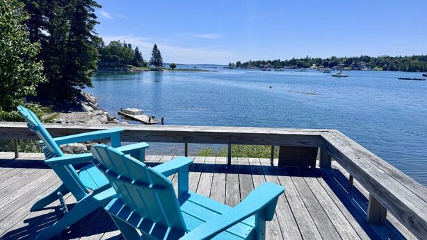 Eine Holzterrasse mit blauen Adirondack-Stühlen mit Blick auf einen ruhigen See mit Booten.