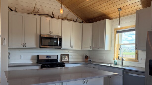 A modern kitchen with white cabinets, a stainless steel microwave, a built-in oven, a large island with stools, and a window with a view of the outdoors.