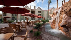 Une terrasse extérieure avec des tables et des chaises, des parasols rouges et une cascade décorative.
