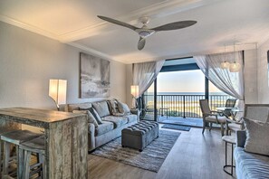 A spacious living area with a large window offering a view of the beach, a ceiling fan, and a bar area. - Beachfront Sand Key Condo w/ Gulf Views (Clearwater)