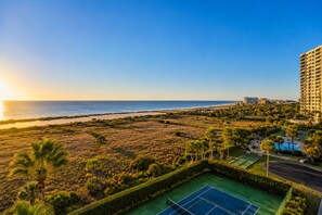 A coastal view with a tennis court, palm trees, and a high-rise building. - Beachfront Sand Key Condo w/ Gulf Views (Clearwater)