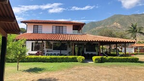 A two-story building with a tiled roof, a covered patio area, and a well-maintained garden with hedges and a small tree. - Peaceful Farmhouse • 10 Min to Roldanillo • Perfect for Paragliders (Bolivar)