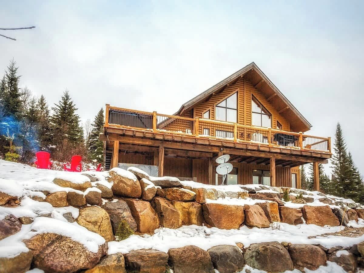 Eine Holzhütte mit Balkon vor einer verschneiten Landschaft mit Bäumen.