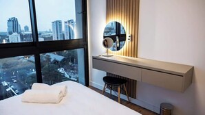 Une chambre d’hôtel moderne dotée d’un grand lit, d’un bureau avec un miroir et offrant une vue sur le paysage urbain grâce à de grandes fenêtres.