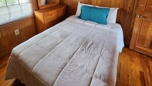 Une chambre avec un plancher en bois, un lit recouvert d’une couette blanche, une table de chevet en bois et une fenêtre avec des stores.