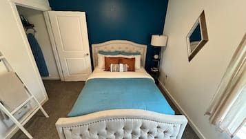 Quarto com cabeceira estofada, cama com colcha azul, porta branca e mesa de cabeceira com abajur.