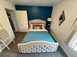 Un dormitorio con cabecera capitonada, cama con cubrecama azul, puerta blanca y mesita de noche con lámpara.