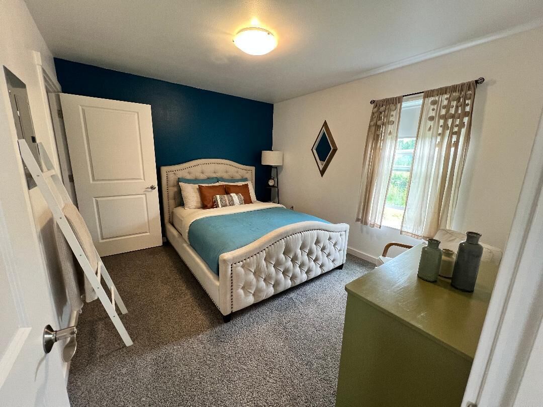 A bedroom with a bed, a desk, a chair, a lamp, and a window with curtains.