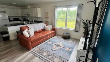 Et moderne køkken med hvide skabe, et spiseområde med en brun lædersofa og et stort vindue med udsigt til grønne omgivelser.
