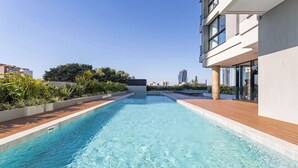 Une piscine dotée d’une terrasse en bois, entourée de verdure et de bâtiments modernes.