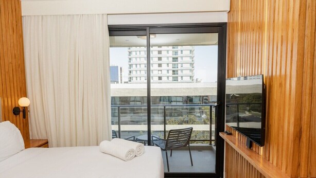 Habitación de hotel con cama, televisor montado en la pared, balcón con silla y vista a edificios.
