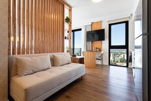 A modern living room with a sofa, a wooden partition, a flat-screen TV, and a city view through large windows. - A luxury space designed to make you feel at home (Manora)