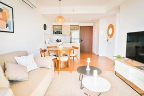 Ein modernes Wohnzimmer mit einer Couch, einem Couchtisch und einem Esstisch mit Stühlen. Im Hintergrund ist eine Küche mit Kühlschrank und Mikrowelle erkennbar.