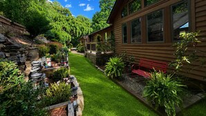 A wooden house with large windows, a stone retaining wall, and a well-maintained garden with a bench. - Five Kings Retreat - 5 BD 3 BTH Mountain Home (Asheville)