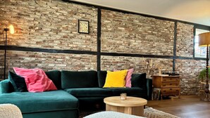 Ein gemütliches Wohnzimmer mit einem grünen Ecksofa, einem runden Holztisch und einer Ziegelwand mit einem gerahmten Bild.