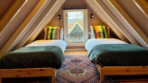Ein kleines, gemütliches Schlafzimmer im Dachgeschoss mit zwei Betten, einem gemusterten Teppich und Blick auf ein Haus durch ein Fenster.