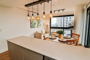 Een moderne keuken met een centraal eiland, hangende verlichting, een zithoek met houten stoelen, een bank en een groot raam met uitzicht op de stad.