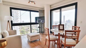 Een moderne woonkamer met een bank, poef, eettafel, stoelen, een televisie en grote ramen met uitzicht op de stad.