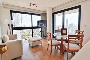 Ein modernes Wohnzimmer mit einer Couch, Hocker, Esstisch, Stühlen, einem Fernseher und großen Fenstern mit Blick auf die Stadt.