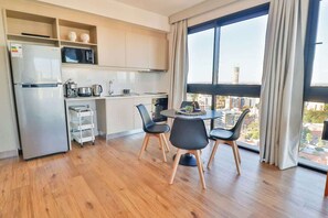 Een moderne keuken met een koelkast, magnetron en koffiezetapparaat, een eethoek met een tafel en stoelen, en een groot raam met uitzicht op gebouwen.