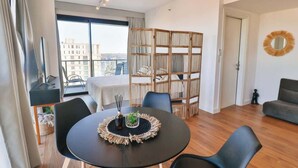 En moderne stue med et rundt bord, sorte stole, en reol og en balkon med udsigt til bygninger.