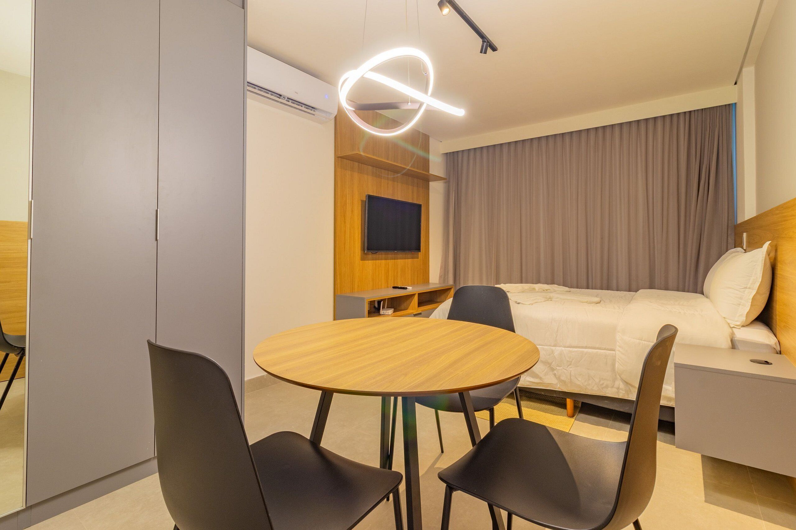 Habitación de hotel con cama, televisión, una mesa redonda de madera con sillas y una lámpara moderna.