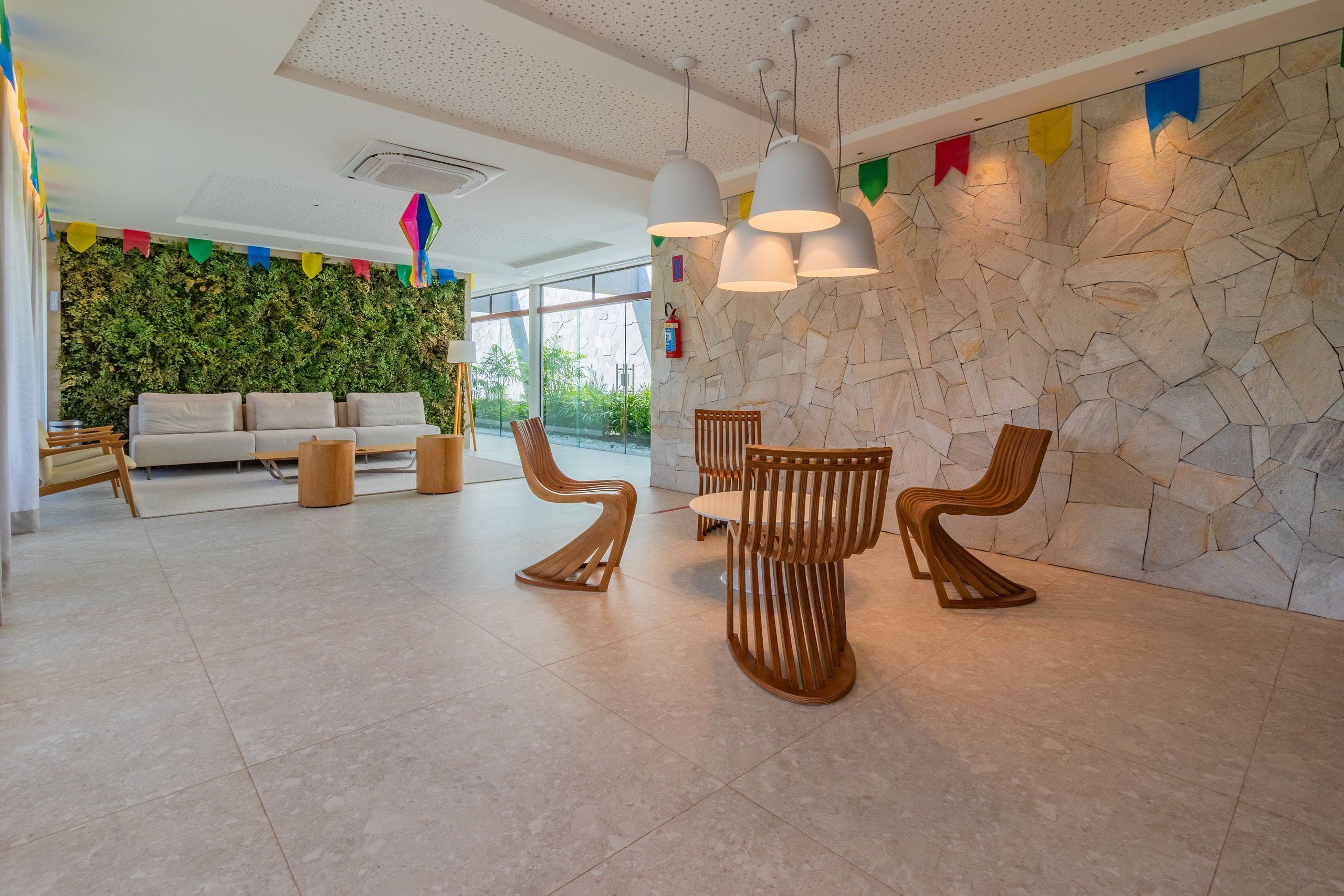 Un vestíbulo moderno con pared de piedra, pared verde con plantas, un sofá, sillas de madera y lámparas colgantes.