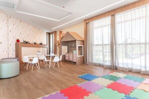Ein moderner Kinderspielraum mit Holzboden, weißen Tischen und Stühlen, einer Spielküche und einer bunten Schaumstoff-Puzzle-Matte.