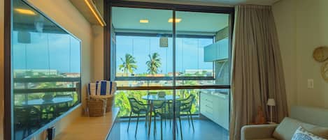 A modern living room with a large window, a sofa, a small table, and a balcony with a view of palm trees.