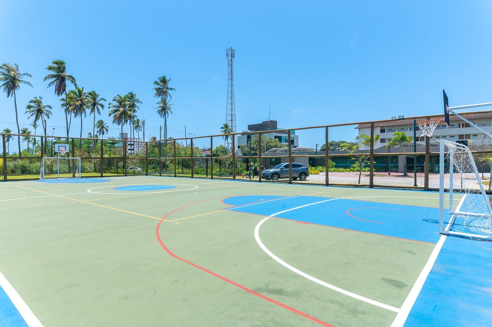 An outdoor basketball court with a fence, a car, and palm trees in the background.