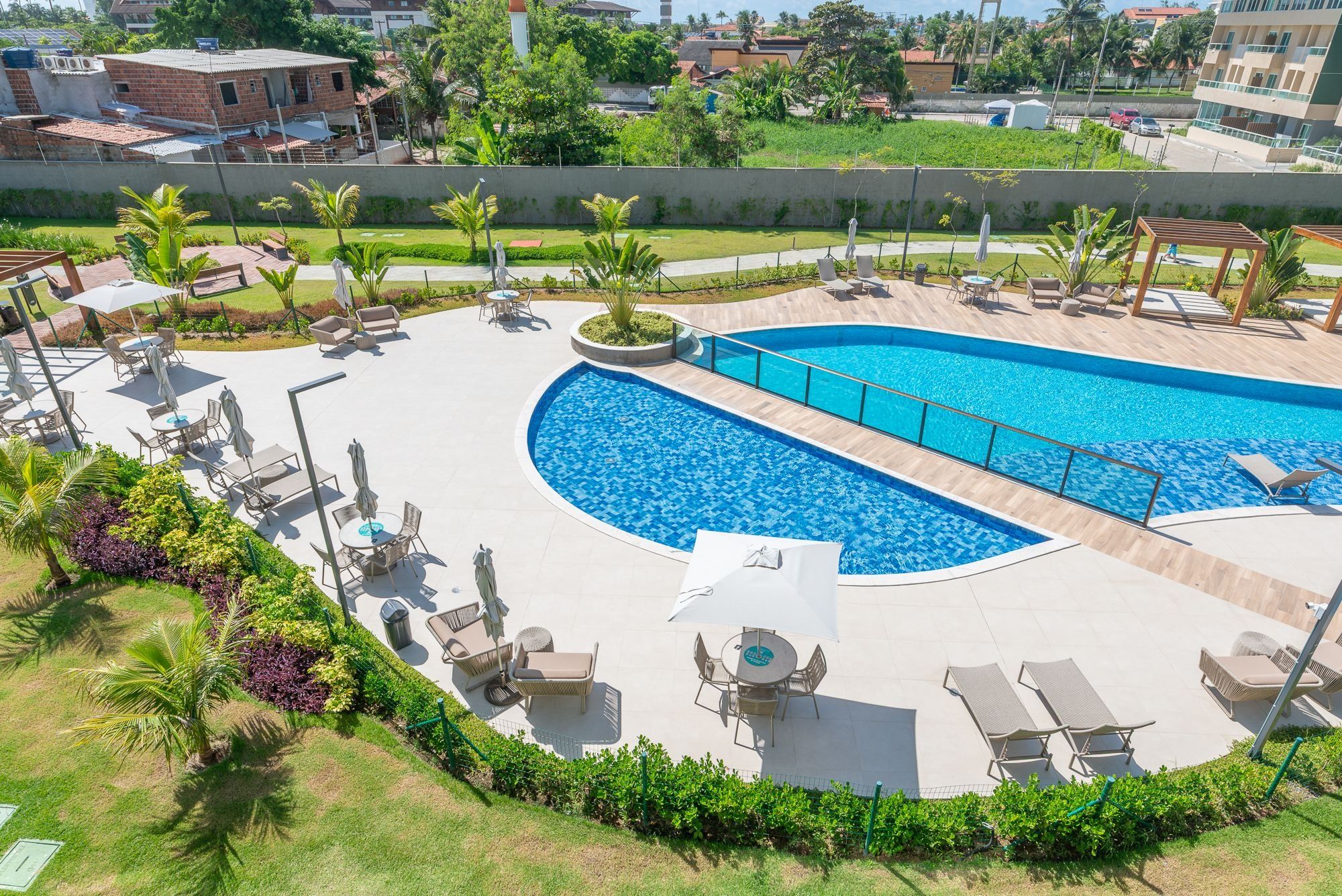 Piscina com espreguiçadeiras e mesas, rodeada por vegetação e vista para a cidade.