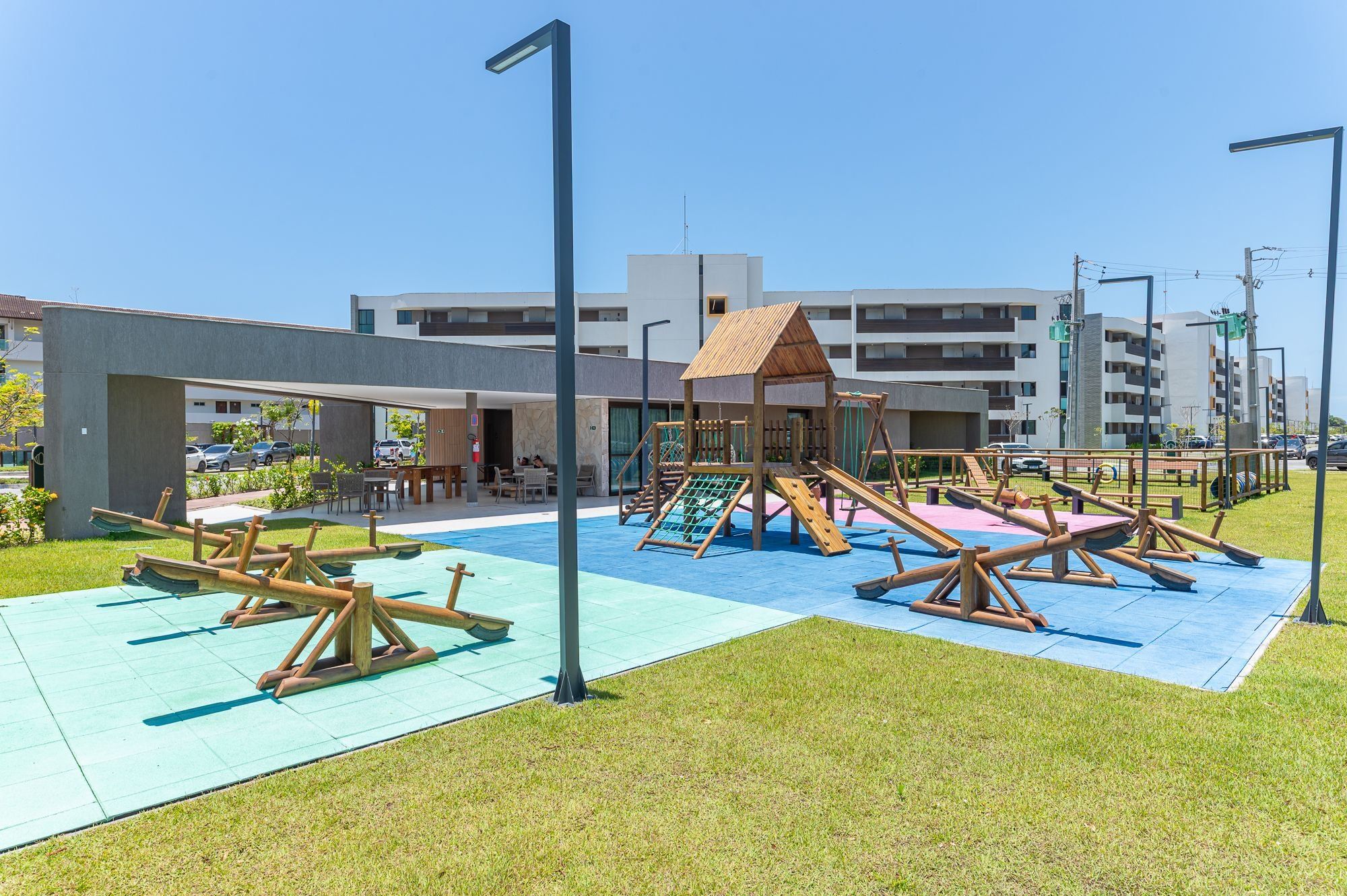 Un parque infantil con columpios y toboganes, una zona de estar techada, y un edificio con balcones al fondo.