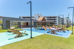 A playground with swings and slides, a covered seating area, and a building with balconies in the background. - Beach Class Summer by Carpediem (Ipojuca)