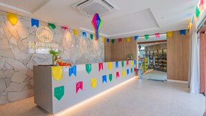 A reception area with a stone wall, a neon sign, a counter, and colorful flags. - Beach Class Summer by Carpediem (Ipojuca)