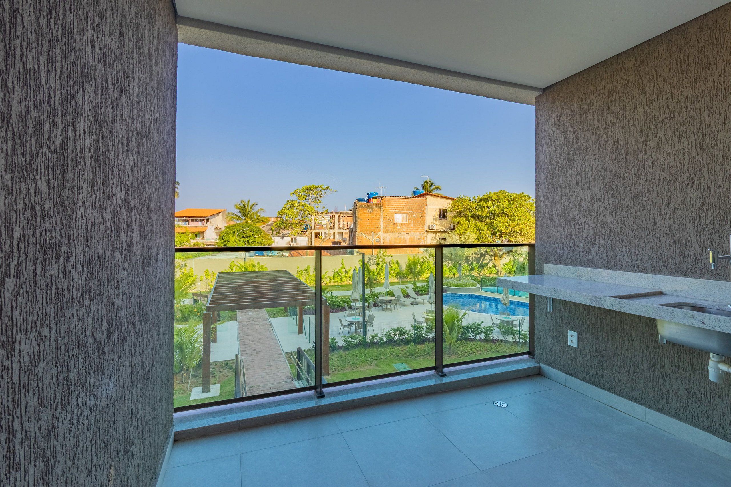 Balcón con vistas a una piscina y vegetación, zona de cocina moderna y suelo de baldosas.